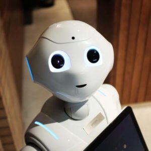 A small humanoid robot with a smooth white exterior and large, expressive black eyes looks up toward the camera with a friendly expression. The robot is holding a tablet or screen against its chest. The background includes a tiled wall and wood-panelled surfaces, suggesting an indoor environment like a tech exhibit or office.